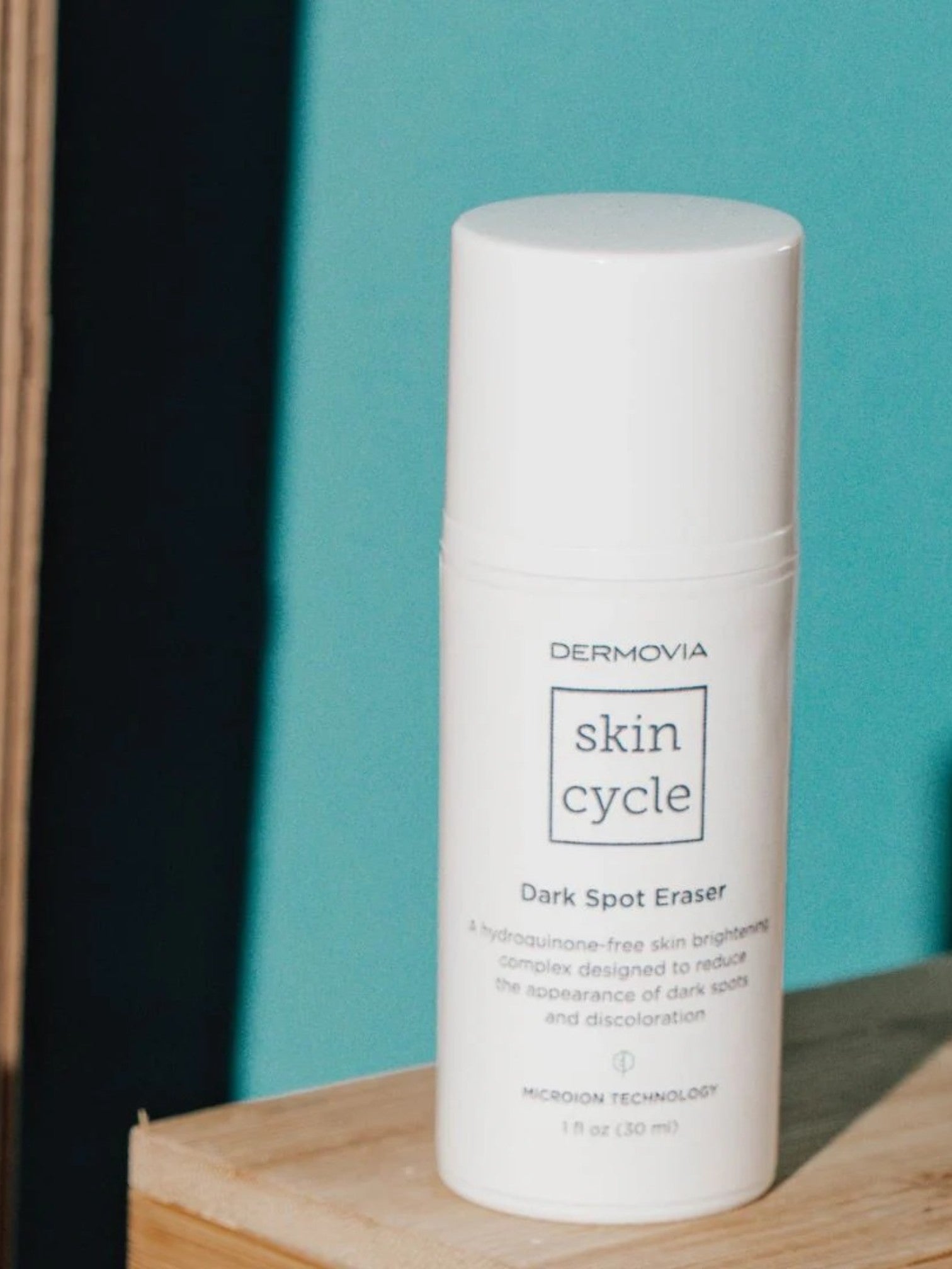 Dermovia Skin Cycle Dark Spot Eraser bottle on a wooden surface with a teal background